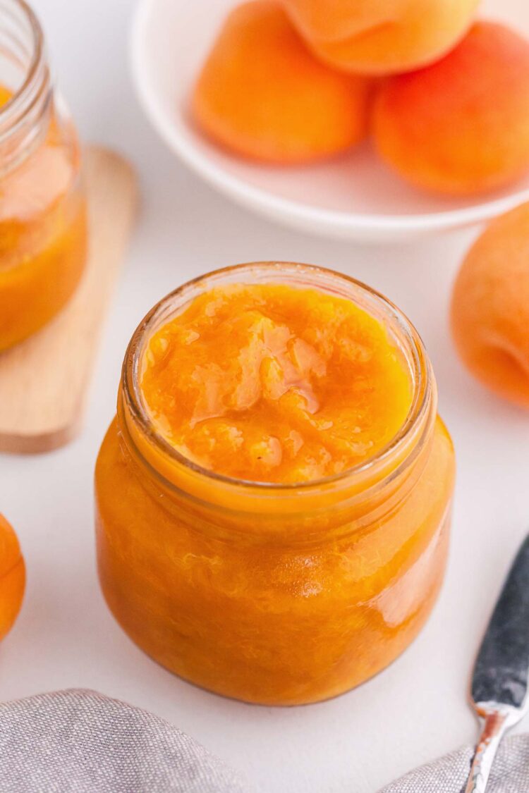 a jar of apricot preserves on a white table with more apricots in the background