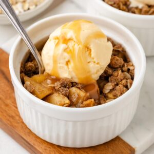 apple crumble in a white ramekin on a cutting board with ice cream and caramel