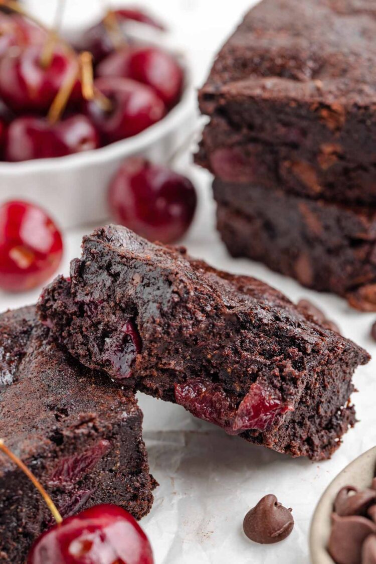 a cherry brownie with a bite taken out of it on a white table with cherries in the background