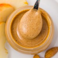 pin image on how to make almond butter with a jar of smooth, golden almond butter in a jar with a spoon dipping into the butter with sliced apples on the side