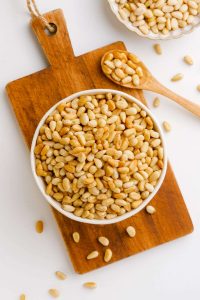 photo of a bowl of toasted pin nuts on a wooden cutting board and a wooden spoon to the side with scattered pine nuts in the background