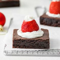 two brownies with Santa hats made of whipped cream and strawberry on a cooling rack