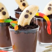 close up photo of healthy chocolate pudding Halloween dirt cups with a banana with a face made out of chocolate chips and gummy worms on top