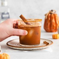 photo of a hand lifting a pumpkin margarita garnished with a cinnamon stick and rimmed in pumpkin spice