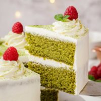 slice of two layered matcha cake with whipped cream cheese frosting with a swirl of frosting on top with a bright red raspberry