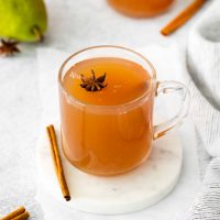 a full mug of pear cider on a white coaster garnished with star anise with a cinnamon stick next to the mug as an accent