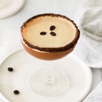 top view image of a vegan espresso martini in a martini glass on a white plate garnished with three roasted coffee beans