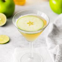 martini glass on a cocktail napkin full of vodka mocktail garnished with sliver of green apple with green apples and limes scattered in background