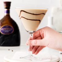 hand holding martini glass stem with Godiva chocolate martini with chocolate swirls and a Godiva liqueur bottle in background