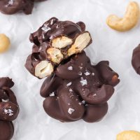 two chocolate covered cashew clusters one with bite taken out both garnished with flaky sea salt with roasted cashews and clusters in background