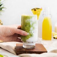 image showing a hand lifting a tall glass of swirled cream and vivid green, pineapple matcha Starbucks copycat drink garnished with a slice of pineapple