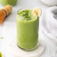 tall glass full of bright green matcha protein shake with a dusting of matcha powder on top and a sliver of banana for garnish on a white marble coaster