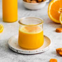 a small glass jar on a rustic white coaster plate full of turmeric ginger shots with orange slices and chunks of turmeric root in the background