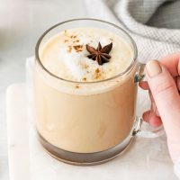 image showing hand reaching for a mug of chai tea latte Starbucks copycat it's foamy and garnished with a star anise and a dash of cinnamon