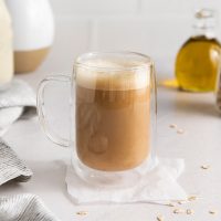 side view of a glass mug of frothy coffee using the olive oil coffee recipe of Starbucks copycat Oleato latte it is golden brown with frothy oat milk layered on top and in the background you see oat milk in a glass jar and olive oil to the side
