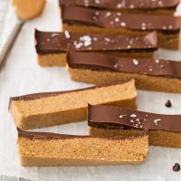 image showing vegan paleo protein bars on white parchment paper with a spoon of almond butter and chocolate chips scattered. The bars are shown top view to see the chocolate top and two are flipped on their side to see the smooth texture of the golden brown bars.