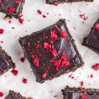 Image of vegan date brownies on a white background with freeze-fried raspberries pieces on top and scattered in the background.