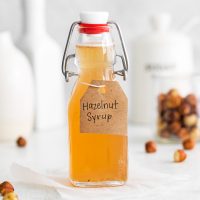 image showing homemade hazelnut coffee syrup in a glass jar with a small glass jar of toasted hazelnuts in the background