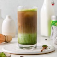 side view image of matcha coffee in a tall glass, layered with green matcha on the bottom and coffee on top with a spoon with matcha tea powder off to the side