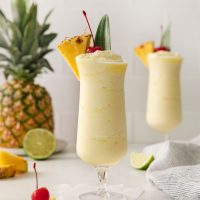 photo of a frozen pineapple daiquiri in a tall glass garnished with a pineapple wedge and a cherry in the background you see a half of a lime and fresh pineapple