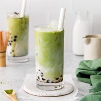 side view photo of a tall glass of green and white layered matcha bubble tea with tapioca pearls at the bottom with another glass of the same in the background
