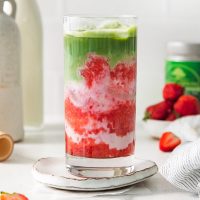 side view image of a tall glass of a strawberry matcha latte with a blended, red strawberry layer at the bottom, a silky layer of milk of choice and vibrant green matcha on top