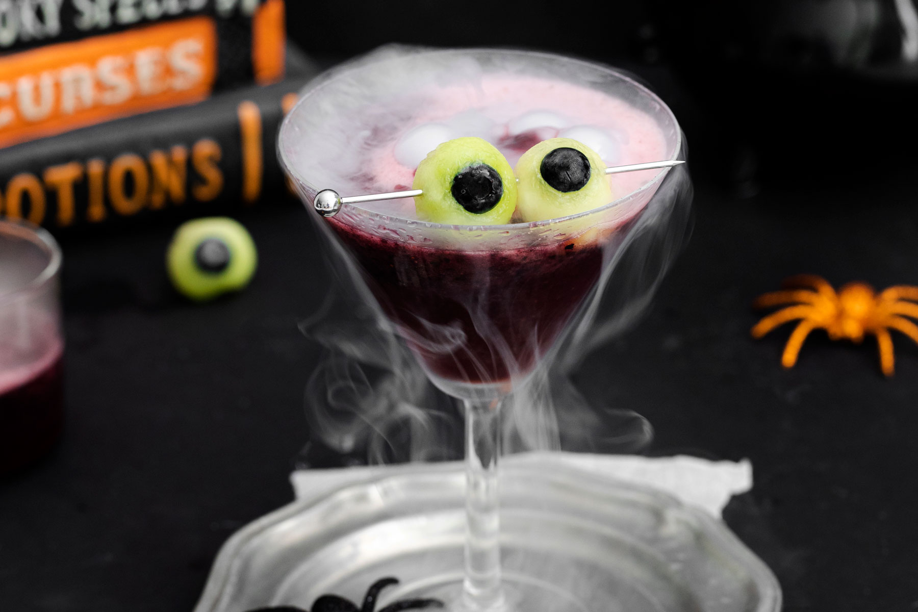A spooky cocktail in a martini glass with wisps of dry ice smoke. The drink is purple, topped with fake eyeballs skewered on a stick. In the background, there are Halloween-themed decorations, including a toy spider and books labeled "Curses" and "Potions.