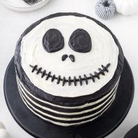 top view photo of a Jack Skellington cake that looks like a skeleton face using black cocoa to get a stark black eye and mouth details on top of a multi-layered chocolate cake with white vanilla cheesecake frosting in between.