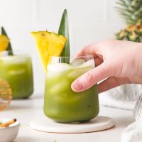 image of a hand picking up a vibrant green matcha cocktail with a pineapple sliver as garnish