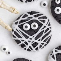 close up photo of a black cocoa mummy cookie decorated with white piped frosting to look like bandages and two candy eyes with skeleton fingers lurking in the background