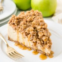 image of a slice of apple crumble cheesecake on a plate with green apples in the background and a fork to the side