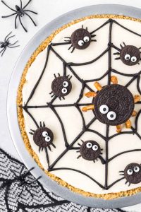 a spider pie showing a round 3-layer pumpkin and vanilla cheesecake decorated with a spider web with one big Oreo mama spider and 8 mini Oreo spider babies