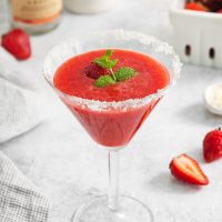 top view of a vibrant, red strawberry martini garnished with a sprig of mint with sliced strawberries in the background