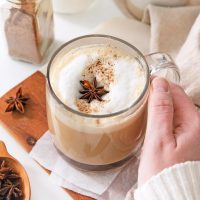 photo of a hand cupping a mug of a dirty chai latte made with chai tea and spice, frothy milk and a shot of espresso and garnished with star anise and a dusting of chai spice