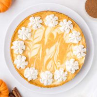 top view photo showing a pumpkin swirl cheesecake which is dusty orange and cream in color with piped whipped cream rosettes around the edge of the cheesecake on a white platter