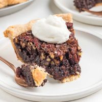 photo of a slice of cocoa pecan pie on a white plate with a fork with the end of the slice on it and a dollop of whipped cream on the pie