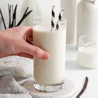 photo of a hand lifting a tall glass of vanilla milk off a coaster with two black and white stripped straws