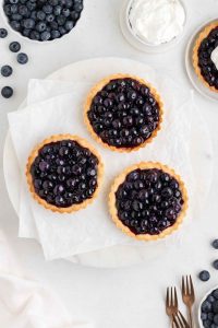 Overhead photo of three blueberry tartlets with golden brown crusts on white parchment paper with several blueberries scattered in the background