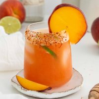 Photo of a peach margarita with a salted rim and garnished with a peach wheel and a slice of lime.