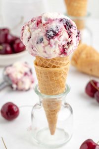 Photo of a black cherry ice cream scoop on a cone with extra cones, scoops of ice cream and fresh cherries in the background