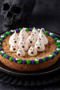 Photo of a Halloween cookie cake with green and purple rim with candy eyeballs for decoration in the middle of the cake are several meringue ghosts making it a perfect Halloween dessert