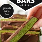 A hand holding a rectangular matcha protein bar, showing a bright green center and a thick chocolate coating on top. In the background, more bars sit on a wire cooling rack. A black circle badge on the left says "gluten & dairy-free."