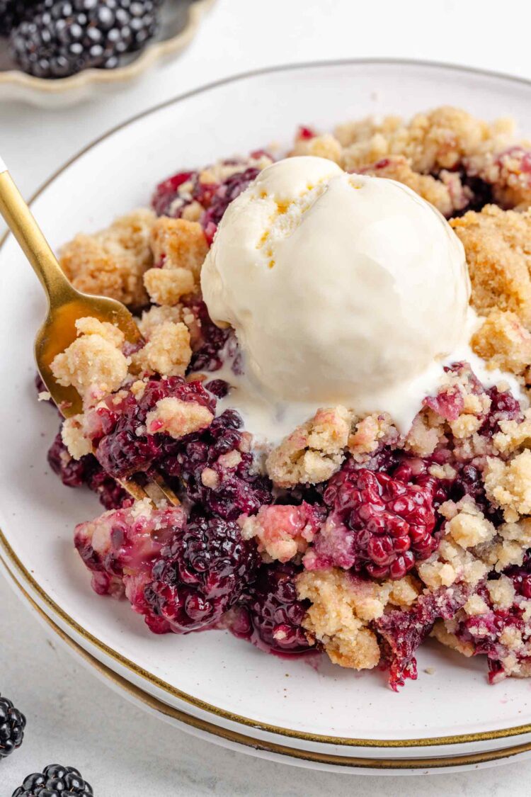 close-up of blackberry crumble on a white plate with a scoop of vanilla ice cream on top and blackberries in background