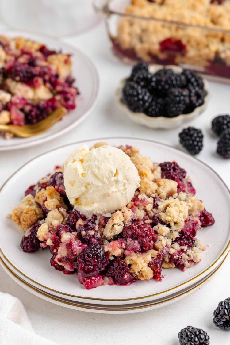 close-up of blackberry crumble on a white plate with a scoop of vanilla ice cream on top and bowl of blackberries in the background