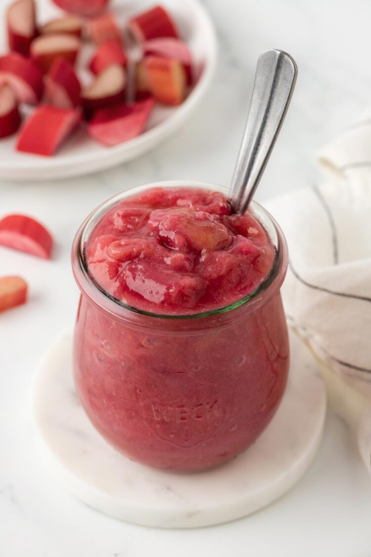 close-up of rhubarb compote in a small glass jar on a white table with a spoon digging into it