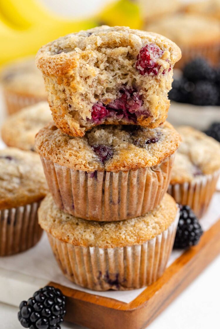 stack of 3 banana blackberry muffins on a cutting board with blackberries in background