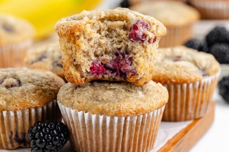 banana blackberry muffins with a bite taken out of the top muffin, sitting on a cutting board