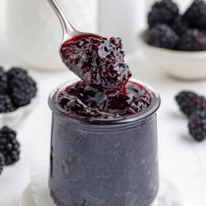 spoon digging into a jar filled with blackberry jam with more blackberries in the background