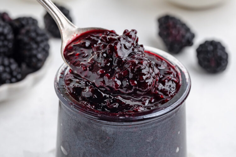 spoon digging into a jar filled with blackberry jam with more blackberries in background