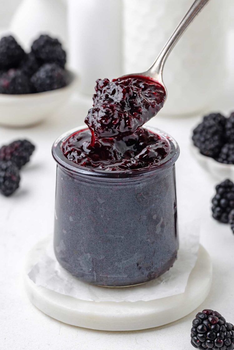 spoon digging into a jar filled with blackberry jam with more blackberries and jug of milk in background
